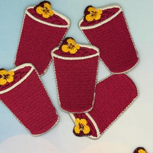 May include: Embroidered patches in the shape of a coffee cup, in a deep red color, with a white border. Each patch features a yellow flower at the top. The patches are arranged on a light blue and white background.