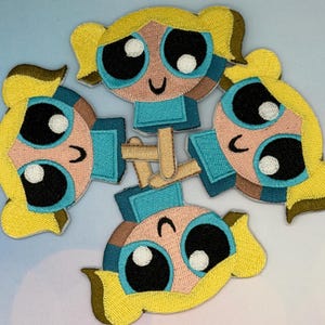 May include: Embroidered patches featuring a cartoon character with blonde pigtails, large blue eyes, and a light blue dress. The patches are in a circular arrangement, showcasing the character's face and torso.