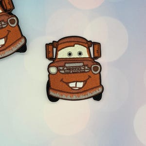 May include: An embroidered iron-on patch featuring a rusty brown tow truck character with a wide, toothy grin, large white eyes, and a silver front bumper. The design includes black outlines and side mirrors, rendered in a cartoon style with thread.