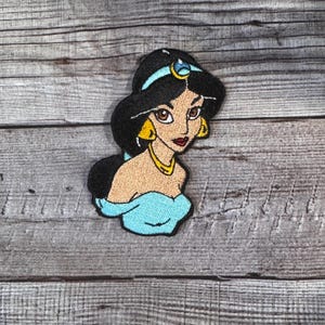 Puede incluir: Parche bordado con una representación de dibujos animados de una princesa con cabello negro, una diadema azul y un vestido turquesa. El parche tiene aretes dorados y un collar dorado. El fondo es una superficie de madera.