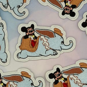 May include: An embroidered patch featuring a light blue elephant with large brown ears and a yellow hat, carrying a black mouse character with a yellow bowtie and red shorts on its back. The design is outlined in white thread on a fabric base.