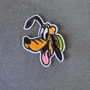 Op de afbeelding: Geborduurde patch met Pluto, de Disney-tekenfilmhond. De patch heeft een witte rand, met Pluto's gezicht in oranje, zwart en wit. Zijn tong is roze en er is een groene vlek op zijn rug zichtbaar.