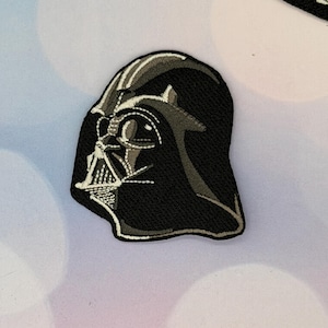 May include: Embroidered patch depicting Darth Vader's helmet profile. The patch is predominantly black with grey and white accents, accurately representing the character's mask. The design is detailed and suitable for application on clothing or accessories.