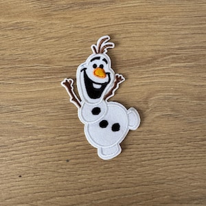 Może przedstawiać: Biała haftowana naszywka do prasowania Olafa, postaci bałwana z filmu Kraina Lodu, z brązowym noskiem z gałązki i czarnymi oczami.
