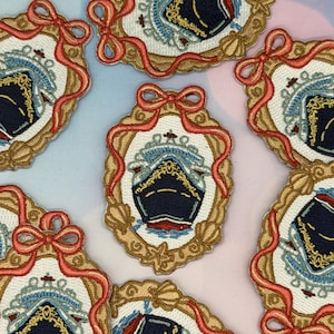Puede incluir: Parches bordados con un diseño de crucero azul marino dentro de un marco ovalado adornado. El marco es dorado con una cinta y un lazo rojos. Se incluyen detalles de conchas marinas y estrellas de mar. Los parches están dispersos sobre un fondo azul claro y rosa.