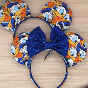 May include: Two headbands with mouse ears. The ears are patterned with Donald Duck's face in yellow, orange, blue, and white. A blue sequined bow is centered on one headband. The headbands are blue.