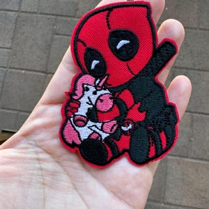 Op de afbeelding: Een geborduurde patch van Deadpool die een roze eenhoorn vasthoudt, in rood en zwart.