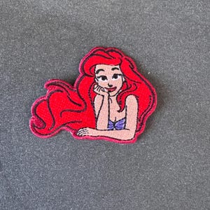 Op de afbeelding: Geborduurde patch met een cartoonillustratie van een zeemeermin met lang rood haar, een paarse top en een glimlachende uitdrukking. De patch heeft een rode rand en een grijze achtergrond.