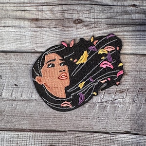 May include: Embroidered patch featuring a woman with long black hair blowing in the wind. The hair is decorated with colorful flowers and leaves. The woman has brown skin and is looking to the right.