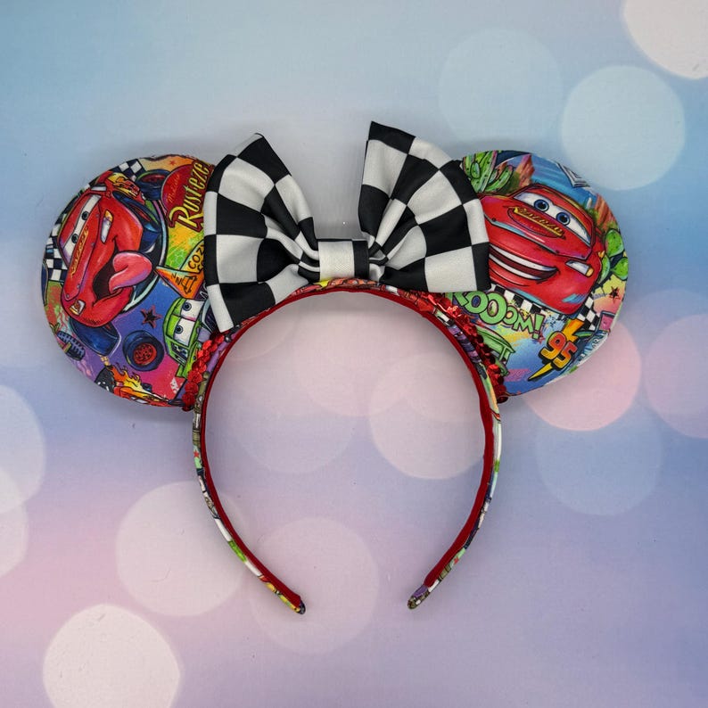 May include: A pair of mouse ears featuring a black and white checkered bow. The ears are decorated with colorful images of Lightning McQueen from the Cars movie. The headband is red.