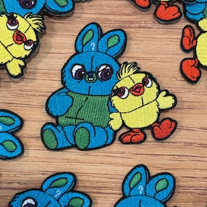 May include: Embroidered patches featuring a blue bunny with a question mark on its ear and a yellow chick. The bunny has purple eyes and a green vest. The chick has red feet and orange sunglasses. The patches are on a wooden surface.