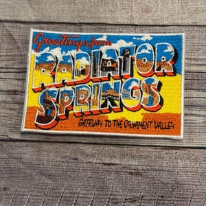 Pode incluir: Um remendo bordado amarelo e azul com o texto "Greetings from Radiator Springs, Gateway to the Ornament Valley".