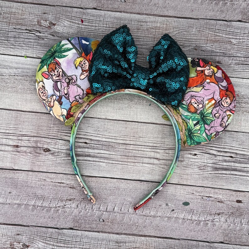 May include: A themed headband featuring two round ears with a colorful Jungle Book print. The headband has a large, sequined teal bow. The ears display scenes with characters from the animated film. The headband is decorated with a floral pattern.