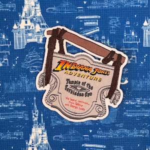 May include: A brown and beige embroidered patch with the text "Indiana Jones Adventure Temple of the Forbidden Eye" and the text "The legend continues... from Disney and George Lucas".