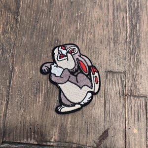 May include: A gray and white embroidered patch of a cartoon rabbit with red ears. The rabbit is looking up and has its mouth open.