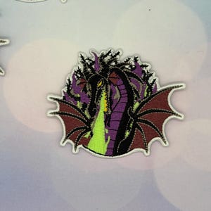 May include: Embroidered patch featuring a dragon with black, purple, and maroon details. The dragon is breathing green flames. The patch has a white border and is likely for clothing or accessories.