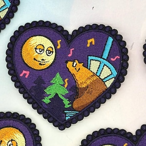 Pode incluir: Um emblema bordado em forma de coração com fundo roxo escuro, apresentando uma lua amarela com um rosto, um urso castanho, um pinheiro verde e uma estrutura azul clara. Notas musicais cor-de-rosa estão espalhadas pelo design, que é contornado por uma borda festonada roxa escura.