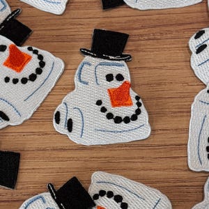Puede incluir: Parches de muñeco de nieve bordados con cuerpo blanco, sombrero de copa negro y nariz de zanahoria naranja. El muñeco de nieve tiene ojos de puntos negros y una boca curva. Los parches están sobre una superficie de madera.