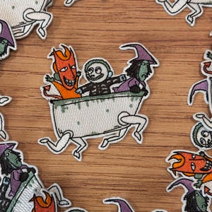 May include: Embroidered patches featuring characters in a bathtub, including a devil, a skeleton, and a witch. The patches have a white border and are set against a wooden background. The characters are in orange, black, purple, and green.