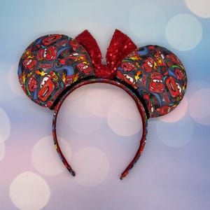 May include: A themed headband featuring two round ears patterned with red race cars on a gray background. A red sequined bow sits between the ears. The headband itself is red with a black trim.