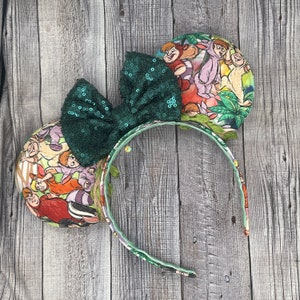 May include: A pair of mouse ears featuring a green sequin bow and round ear designs with a colorful cartoon print. The headband is decorated with leaves and a matching print.