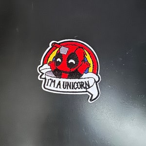 Op de afbeelding: Rode en zwarte geborduurde patch van een cartoon Deadpool-personage met een regenboog erachter en een wit banner met de tekst "Ik ben een eenhoorn".