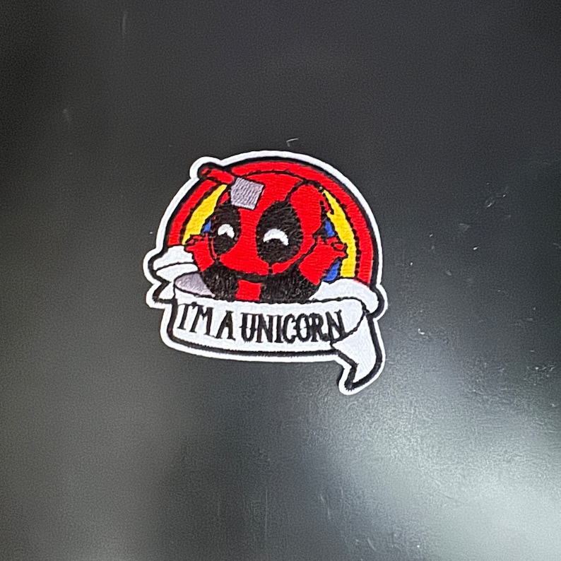 K&ouml;nnte beinhalten: Roter und schwarzer gestickter Aufn&auml;her mit einem Cartoon-Deadpool-Charakter, einem Regenbogen dahinter und einem wei&szlig;en Banner mit der Aufschrift "Ich bin ein Einhorn".