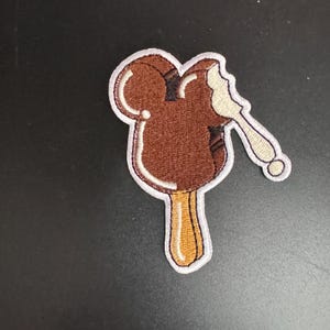 Puede incluir: Un parche bordado con forma de helado de Mickey Mouse con un mordisco, en marrón y blanco.