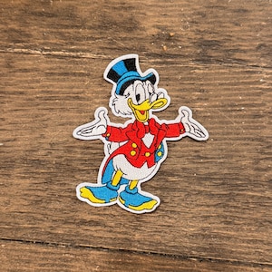 May include: Embroidered patch featuring a cartoon character in a red jacket, blue pants, and a top hat. The character has a yellow beak and is smiling with its arms outstretched. The patch is white around the edges and is on a wooden surface.