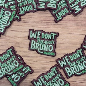 May include: A collection of embroidered patches. Each patch features the phrase "We Don't Talk About Bruno" in green and burgundy. The patches are rectangular with rounded corners and have the text "No No No".