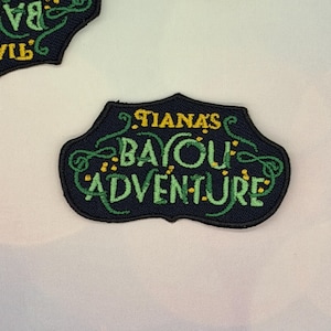 May include: Embroidered patch with the text "Tiana's Bayou Adventure" in green and yellow on a navy blue background. The patch has a decorative border and is likely for clothing or accessories.