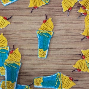 May include: Embroidered patches featuring a yellow ice cream sundae with a cherry on top, set against a blue cup. The patches are scattered on a wooden surface, showcasing the detailed stitching and vibrant colors.
