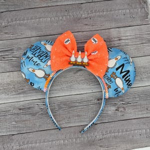 May include: A pair of blue and white mouse ears with a bright orange bow and a small charm of three seagulls. The ears are decorated with the text "Mine Mine Mine" and seagulls.