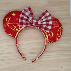 May include: A red headband with mouse ears and a bow. The ears feature a yellow paisley pattern. The bow has a red and white gingham design. The headband is trimmed with gold sequins.