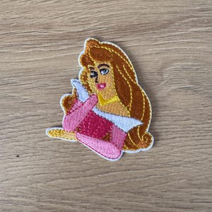 May include: Embroidered patch featuring a cartoon princess in a pink dress and white undergarments. The princess has long, flowing auburn hair and a yellow necklace. The patch is outlined in gold thread and is likely for clothing or accessories.