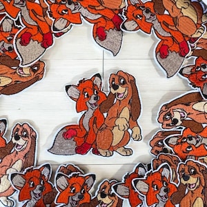 Op de afbeelding: Geborduurde patches met de vos Tod en de hond Copper uit de animatiefilm "Frank en Frey". De patches zijn voornamelijk oranje, bruin en beige, met gedetailleerde steken. De centrale patch toont de twee personages samen.