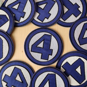 Może przedstawiać: Okrągłe, niebiesko-białe, haftowane naszywki z numerem 4 pośrodku. Naszywki mają niebieską obwódkę i białe tło dla numeru. Naszywki są ułożone na jasnobrązowej powierzchni.