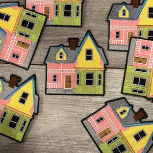 May include: Embroidered patches featuring colorful houses with yellow, pink, and green facades. Each patch has a black border and depicts a house with windows, doors, and a chimney. The patches are arranged on a wooden surface.