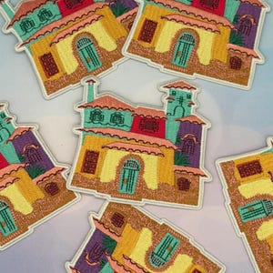 May include: Embroidered patches featuring colorful, stylized houses with arched doorways, teal doors, and red, yellow, and purple roofs. The patches have a white border and are scattered across a light blue background. The patches are likely for clothing or accessories.