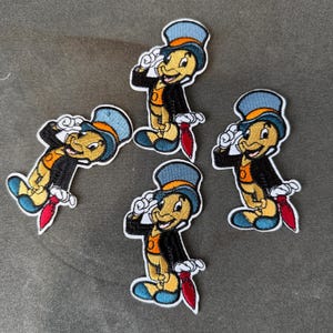 Könnte beinhalten: Vier aufgestickte Aufnäher mit einer Zeichentrickfigur, einer Grille. Die Grille ist gelb mit einem blauen Zylinder, einer schwarzen Jacke, einer orangefarbenen Weste und einer roten Krawatte. Die Aufnäher sind weiß umrandet.