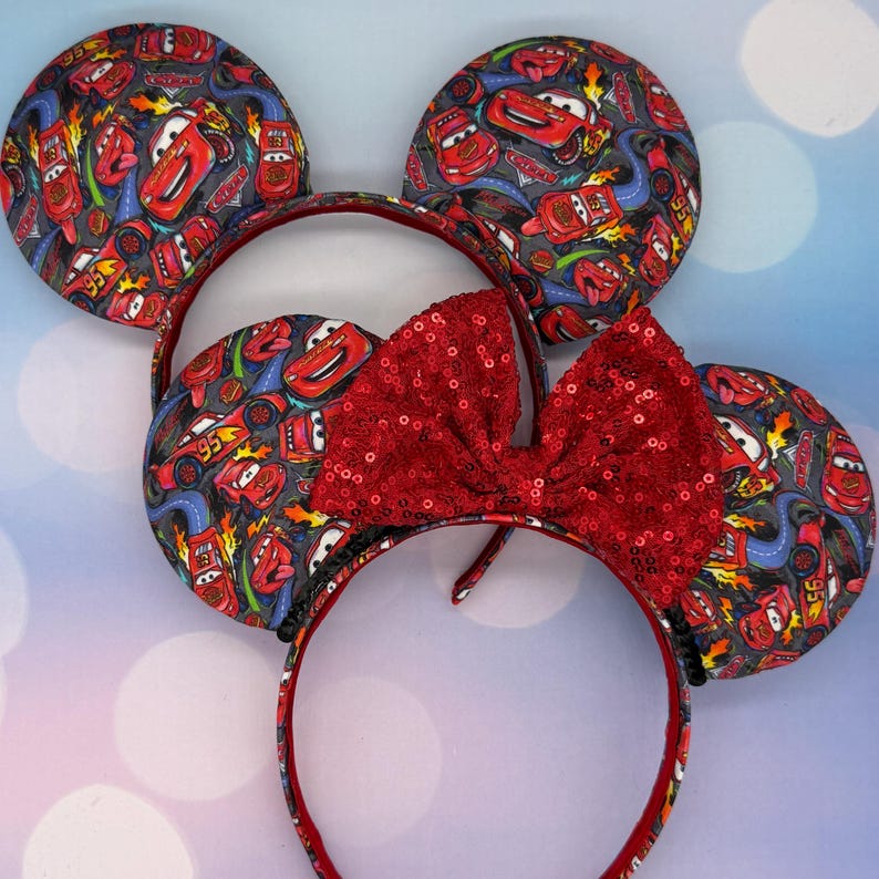 May include: Minnie Mouse ear headband featuring a red sequined bow and ears patterned with the character Lightning McQueen from the movie Cars. The ears and headband are red and black. The background is a light blue.