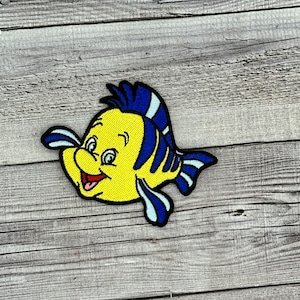 Op de afbeelding: Een geborduurde strijk-op-patch van een cartoonvis met een glimlachend gezicht. De vis is geel met blauwe vinnen en een blauw-wit gestreepte staart.