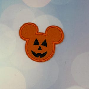 May include: An orange embroidered patch in the shape of a mouse head with a jack-o'-lantern face. The patch features two circular ears, a central round head, and a black stitched design depicting two triangular eyes, a nose, and a wide grin.