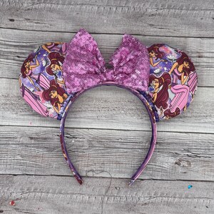 K&ouml;nnte beinhalten: Ein lila Stirnband mit M&auml;useohren. Die Ohren sind mit einem rosa-lila Muster mit einer Zeichentrickfigur verziert. Eine gro&szlig;e, paillettenbesetzte Schleife in passendem Lila sitzt oben auf dem Stirnband.