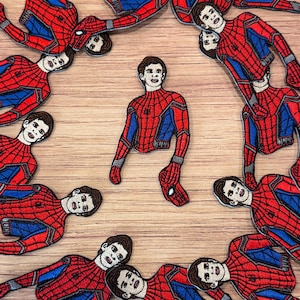 May include: Embroidered patches featuring a red and blue Spider-Man costume design. The patches depict the character in various poses, with a detailed face and web pattern. The patches are arranged on a light brown wooden surface.