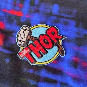 May include: A circular patch featuring the word "THOR" in red, with a hammer graphic. The patch has a light blue background with a yellow border and the words "THE MIGHTY" in red. The background is a mix of blue, red, and black.