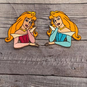 Puede incluir: Dos parches bordados para planchar de La Bella Durmiente, uno en rosa y otro en azul, con una corona amarilla y cabello rubio.