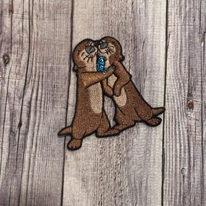 May include: Embroidered patch featuring two brown otters hugging, with one holding a blue object. The otters have sunglasses and are set against a light brown background. The patch is likely for clothing or accessories.