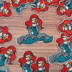 Op de afbeelding: Geborduurde patches met een stripfiguur met rood haar, een turquoise jurk en een zeester. De patches zitten in een zittende pose, met een glimlachende uitdrukking. De patches liggen op een lichtbruin houten oppervlak.