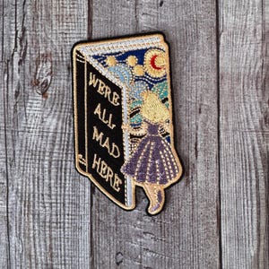 May include: A black and white embroidered patch with a book-shaped design. The book is open and a figure in a purple dress is stepping out of the book. The text on the book reads "We're All Mad Here".